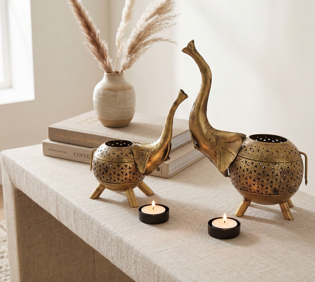 Two brass elephant-shaped candle holders with laser-cut star patterns sit on a beige tablecloth-covered table. One is open and the other is closed with a tealight candle inside, styled with books, a vase of pampas grass, and small tea lights.