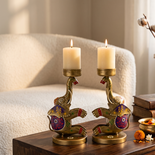 Two ornate brass elephant-shaped candle holders with colorful painted details, each holding a lit cream pillar candle, displayed on a dark wooden table in a cozy living room setting.