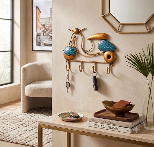 A decorative wall-mounted key rack shaped like a stylized scooter in gold and turquoise blue, shown hanging in a modern entryway with keys, a mirror, chair, and console table.