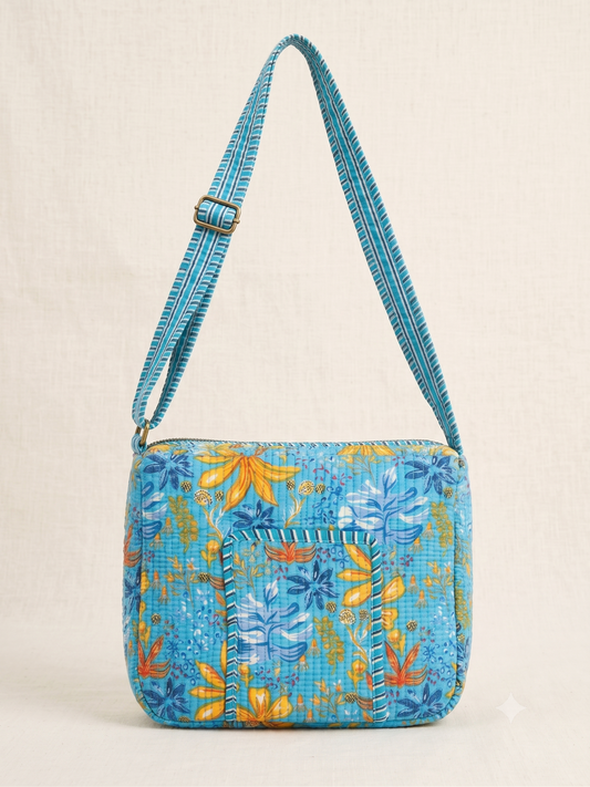 A small crossbody bag with a vibrant blue floral and botanical pattern, featuring a matching patterned shoulder strap with a gold-tone buckle, set against a neutral cream background.